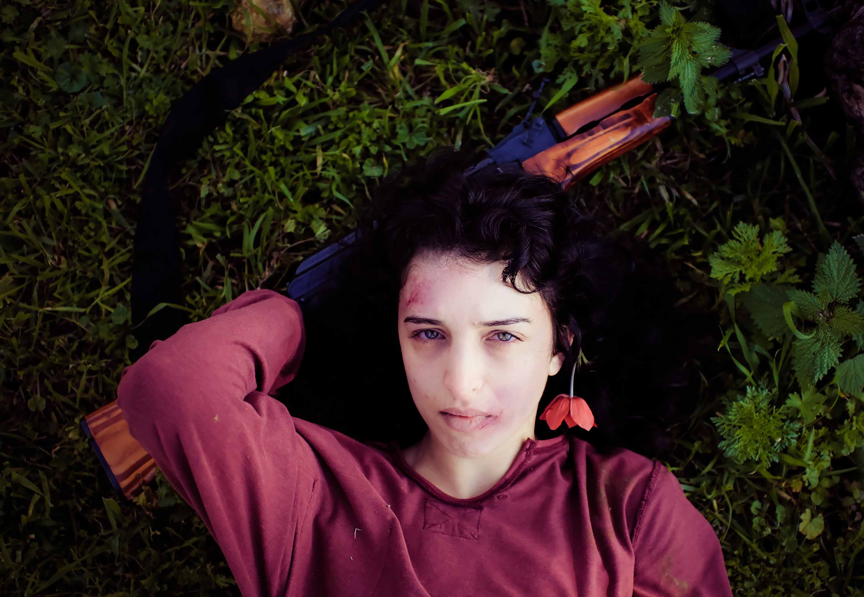  Khawla Ibraheem wears a burgundy long sleeve top while lying on grass. Her head rests on a rifle, and a red flower is tucked behind her ear. She has a bruise on her lip and her forehead.