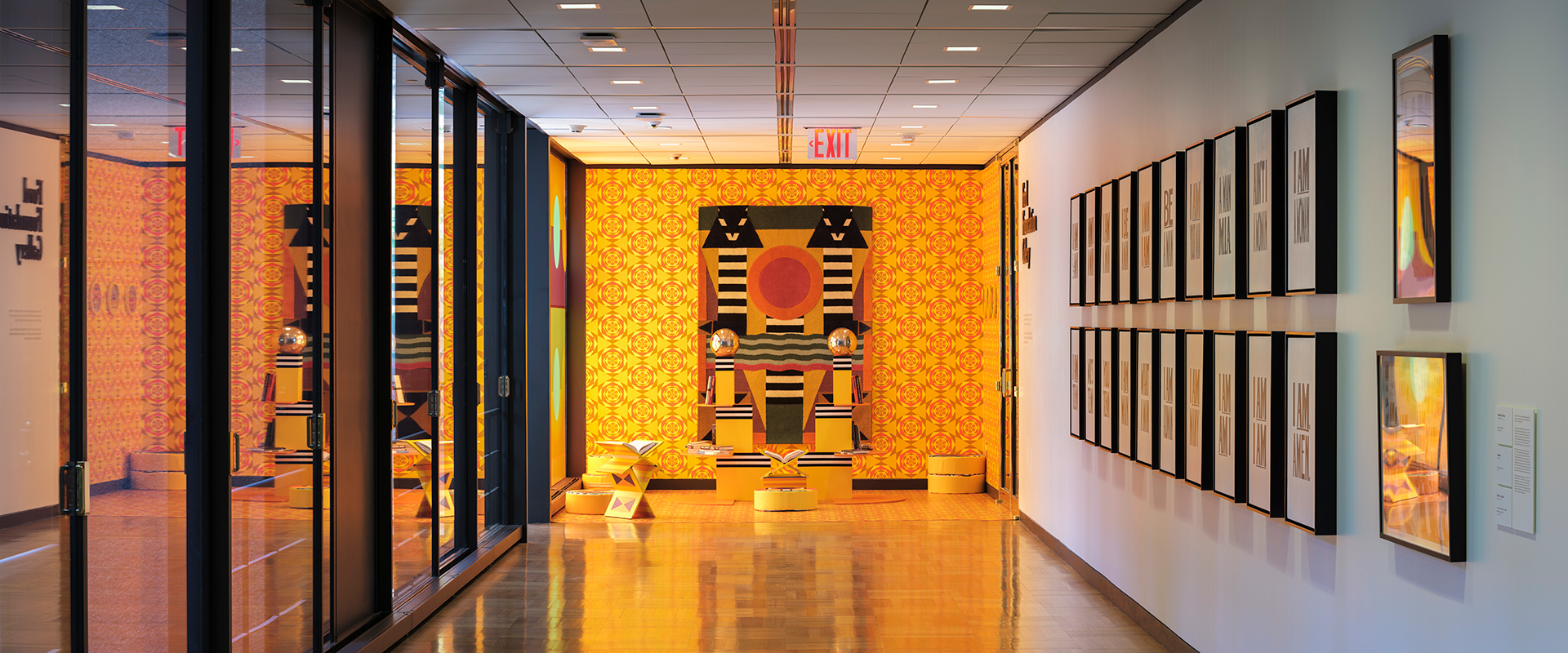 An installation on two walls, a large window, and a section of floor at the end of a hallway. There are wallpapered elements in geometric patterns on all surfaces. A rug hangs vertically on the back wall and there are two sculpted pillars on the floor at waist height.