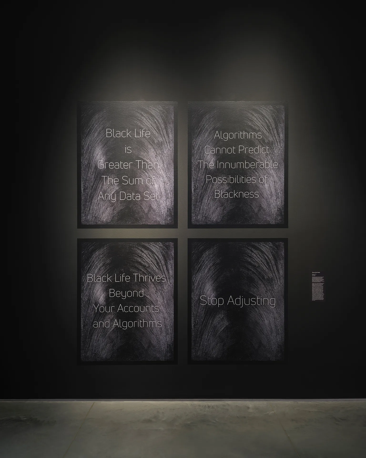 Four posters on painted black backgrounds with visible black brushstrokes. The posters read “Black Life is Greater Than The Sum of Any Data Set,” “Algorithms Cannot Predict The Innumerable Possibilities of Blackness,” “Black Life Thrives Beyond Your Accounts and Algorithms,” and “Stop Adjusting.”