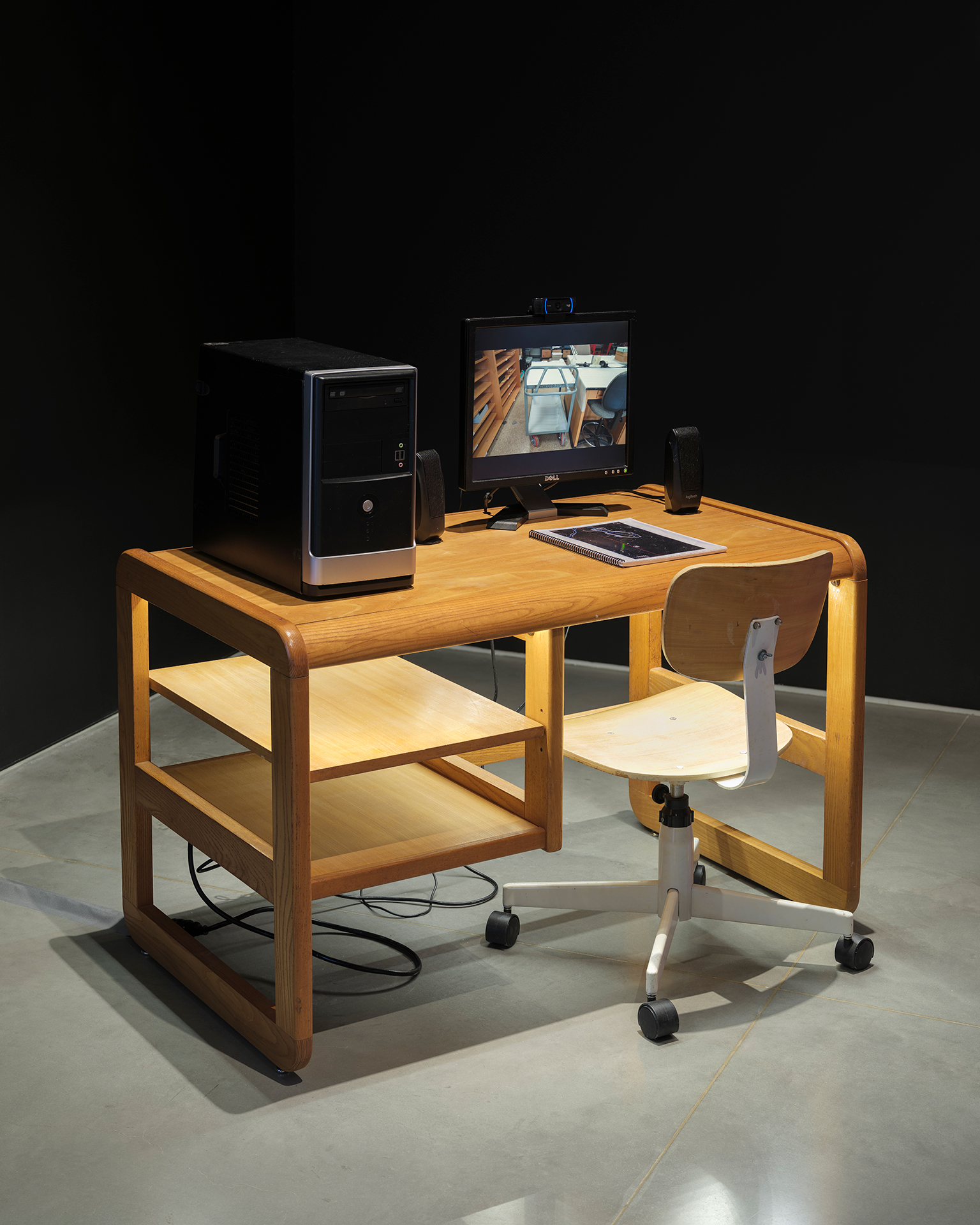 A wooden desk and matching chair holding a computer with monitor and speakers, as well as a spiral bound packet of paper. The monitor shows an image of an archive.