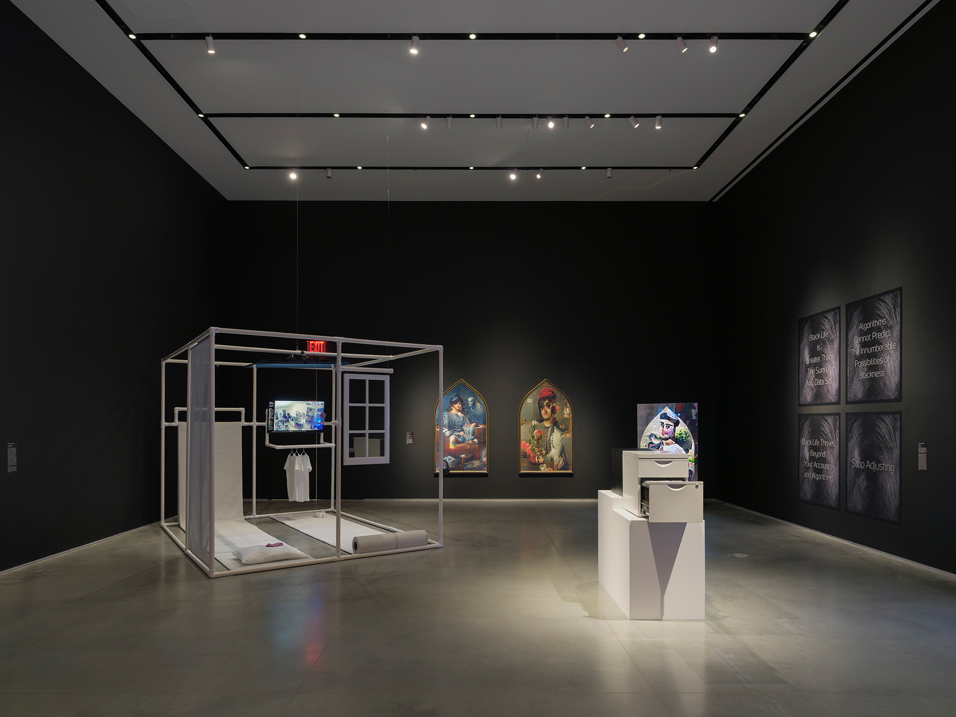 Gallery interior with black walls and a gray floor. There are artworks mounted on the back and left walls and two freestanding artworks in the center of the floor.