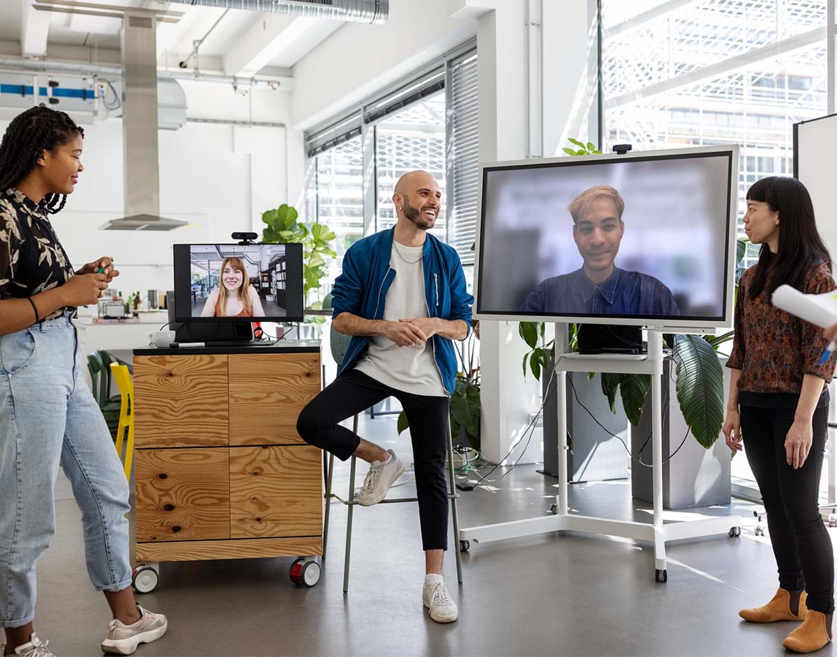 A diverse group of men and women meeting in a hybrid workplace. This group is having video call and meeting with colleagues working remotely. The office space is mostly open floor and brightly lit.