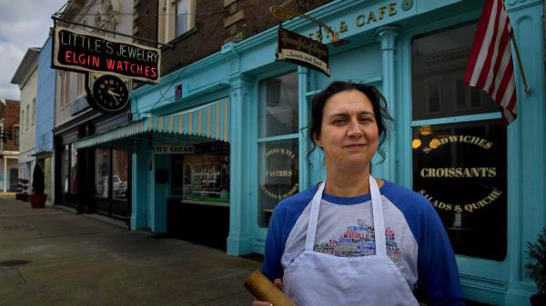 Danielle King, the owner of the Spoonful of Sugar bakery and coffee shop standing in front of her shop. She is a white woman wearing a navy blue shirt with a white apron. 