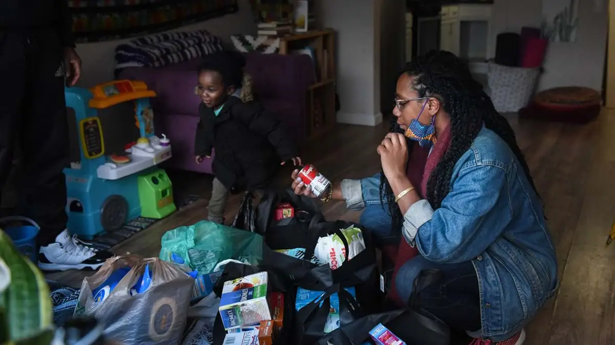 A Black woman is wearing a blue denim shirt with hair braids sitting on the floor looking at a can of food, and a black toddler is walking towards his toys.