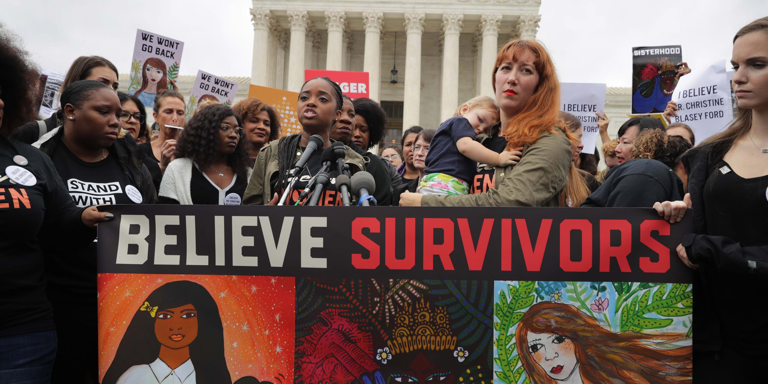 A crowd of women stands behind a banner that reads "Believe Survivors". A Black woman speaks at a podium with an assortment of mics.  A white woman with red hair holding a small child stands next to the podium. 